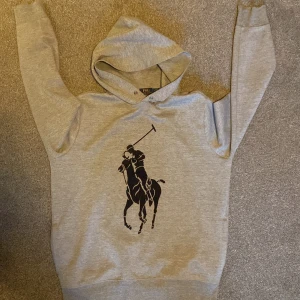 Grå hoodie från Polo Ralph Lauren - Säljer en stilren grå hoodie från Polo Ralph Lauren med det ikoniska polospelarmotivet på framsidan. Tröjan har en bekväm passform och är perfekt för en avslappnad look. Den är tillverkad i mjukt material och har en klassisk huva. Inga skador och sitter perfekt ✅⚡️