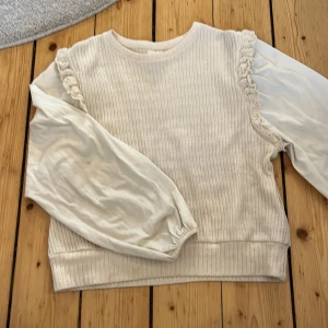 Vit ribbad tröja med volangdetaljer - Säljer en söt vit tröja med ribbad struktur och volangdetaljer vid axlarna. Tröjan har långa ärmar med elastiska muddar och en bekväm passform. Perfekt för en stilren look!