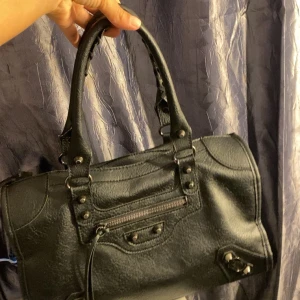 Svart handväska med dragkedja - Snygg svart handväska i skinn med coola detaljer och dragkedja. Säljer på grund av att använder inte längre köpt för 1200 från fyndiq. Den har en praktisk storlek och är lätt att matcha med olika outfits.