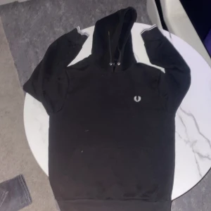 Svart hoodie från Fred Perry - Säljer en stilren svart hoodie från Fred Perry med deras ikoniska logga på bröstet. Tröjan har långa ärmar och en klassisk huva. Perfekt för en avslappnad stil.