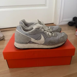 Grå Nike sneakers - Säljer ett par grå Nike sneakers med vit swoosh och snörning. Skorna har en klassisk design med en kombination av syntet och mocka material.  De är använda men ändå acceptabla då stilen inte heller är så stilren 