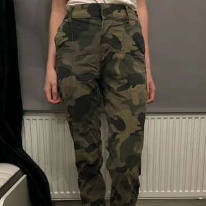 Camouflage cargobyxor - Snygga camouflage cargobyxor med hög midja och flera praktiska fickor. Perfekta för en avslappnad och trendig stil. Byxorna har en klassisk cargodesign med knapp och dragkedja framtill.