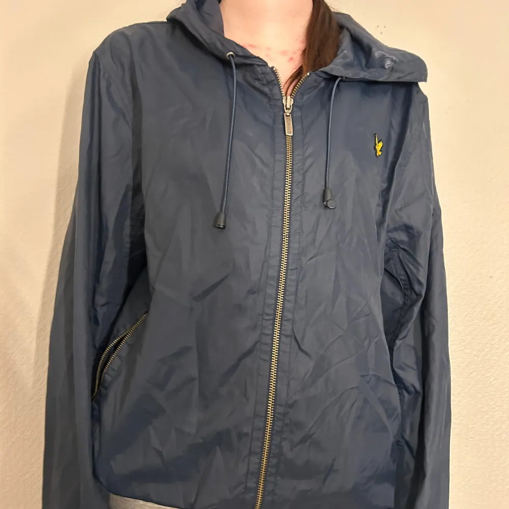 Säljer en snygg blå vindjacka från Lyle & Scott med dragkedja och huva. Jackan har ett gult broderat emblem på bröstet och justerbara snören vid huvan. Perfekt för blåsiga dagar!. Takit.