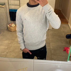 Grå tröja från Tommy Hilfiger - Säljer en stilren grå tröja från Tommy Hilfiger i premium bomull. Tröjan har en liten logotyp på bröstet och ribbade muddar vid ärmslut och nederkant. 