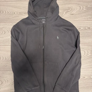 Svart hoodie från Polo Ralph Lauren - Säljer en stilren svart hoodie från Polo Ralph Lauren. Den har en dragkedja framtill och en liten broderad logotyp på bröstet. Perfekt för en avslappnad look.