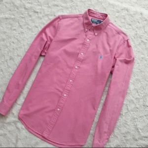 Ralph Lauren Skjorta - Hej! Säljer en rosa skjorta i storlek M, med button-down-krage och blå broderad logga på bröstet. Utmärkt skick utan tecken på användning. Perfekt för både vardag och formella tillfällen.  Hör av dig vid eventuella frågor!