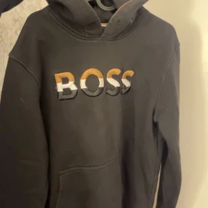 Svart hoodie från Boss - Säljer en svart hoodie från Boss med färgglatt broderat märke på framsidan. Tröjan har en klassisk design med huva och långa ärmar.❤️Pris kan diskuteras