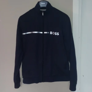 Mörkblå cardigan från Boss - Snygg mörkblå tröja från Boss med dragkedja och stilren design. Jackan har en silverfärgad logotyp på bröstet och en randdetalj vid kragen. Tröjan är storlek s. 