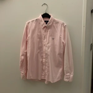 Ljusrosa skjorta från Gant - Säljer en stilren ljusrosa skjorta från Gant i storlek 158/164 cm, perfekt för tonåringar. Skjortan har långa ärmar och klassisk krage med knappar framtill. Den är tillverkad i ett mjukt och bekvämt material.