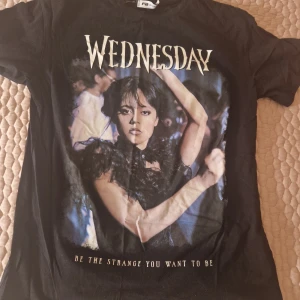Svart t-shirt med Wednesday-motiv från FB Sister - Cool svart t-shirt från FB Sister med ett tryck av Wednesday och texten 'Be the strange you want to be'. Perfekt för fans av den ikoniska karaktären. Kortärmad och i mjukt material.