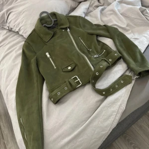 Olivgrön mockajacka från Zara - Snygg olivgrön mockajacka från Zara med dragkedja och bälte i midjan. Jackan har långa ärmar och flera fickor med dragkedjor. Perfekt för en stilren look.
