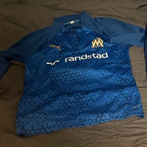Blå Marseille tröja med märker Puma - Snygg blå fotbollströja från Puma med guldiga detaljer och ett mönster av små trianglar. Tröjan har korta ärmar och klubbens emblem på bröstet. Perfekt för fotbollsfans!