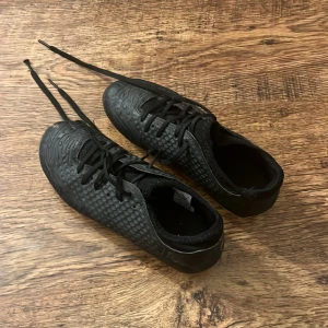 Svarta fotbollsskor med snörning - Snygga svarta fotbollsskor med snörning och mönstrad yta. Skorna har en vit sula med dobbar för bra grepp på planen. Perfekta för fotbollsträning eller match.