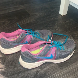 Grå och rosa Nike sneakers - Säljer ett par grå och rosa Nike sneakers i storlek dam 40. Skorna har en snygg kombination av grått och rosa med blå snörning och en vit sula. Perfekta för träning eller vardagsbruk. De är i välanvänt skick men fortfarande bekväma och funktionella.