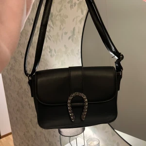 svart handväska - Säljer en stilren svart axelväska i skinn med ett snyggt höstsko spänne framtill. Väskan har en enkel och elegant design, perfekt för både vardag och fest. Den har en justerbar axelrem 💗