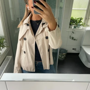 Beige trenchcoat med knappar - Säljer en snygg beige trenchcoat med stora svarta knappar. Jackan har en klassisk design med krage och är perfekt för höst och vår. Den är långärmad och har en normal passform. 