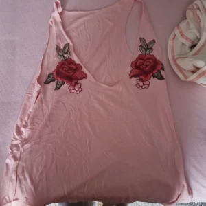 Rosa ärmlös topp med rosbroderi - Säljer en superfin rosa ärmlös topp med v-ringning. Den har vackra broderade rosor i rött och grönt på framsidan. Perfekt för sommardagar eller en avslappnad fest. Toppen är i mjukt material och har en lös passform.