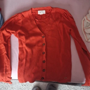 Orange ribbad kofta från Vila - Säljer en snygg orange kofta från lindex. Den är ribbad och har en v-ringad design med knappar framtill. Perfekt för höst och vår, och den har långa ärmar som håller dig varm. Superfin att ha över en topp eller klänning!