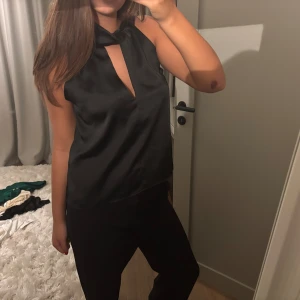 Svart ärmlös topp med knyt - Säljer en elegant svart ärmlös topp med en snygg knytning vid halsen. Toppen har en stilren design med en liten öppning framtill, perfekt för både fest och vardag. Materialet ser ut att vara mjukt och glansigt, vilket ger en lyxig känsla.