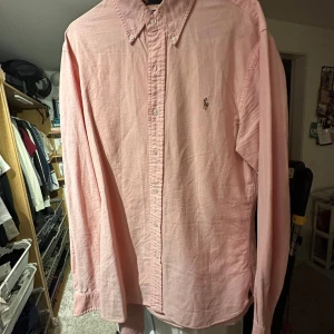 Rosa skjorta från Polo Ralph Lauren - Säljer en snygg rosa skjorta från Polo Ralph Lauren i storlek M. Skjortan är slim fit och har långa ärmar. Den är i bomull och har klassiska knappar framtill. Perfekt för både vardag och fest!
