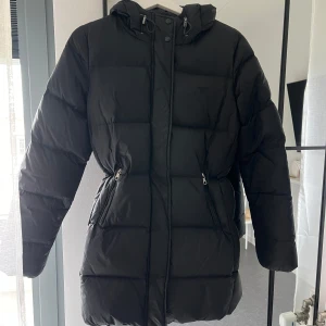 Svart dunjacka från GANT - Säljer en snygg och varm svart dunjacka från GANT. Jackan har dragkedja och knappar framtill. Den är perfekt för kalla vinterdagar och har en huva. Säljer även likadan i kortare modell! 