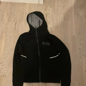 Svart huvtröja från Hugo Boss - Snygg svart huvtröja från Hugo Boss med dragkedja och vit logga på bröstet. Tröjan har en grå insida i huvan och vita detaljer vid fickorna. Perfekt för en casual look.