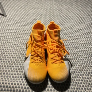 Orange fotbollsskor från Nike - Säljer ett par orangea fotbollsskor från Nike med snörning och svarta detaljer. Skorna har en vit sula med dobbar, perfekt för spel på gräs. De har en sportig design med en blixtdetalj på sidan.