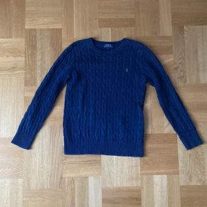 Blå stickad tröja från Polo Ralph Lauren - Säljer en snygg blå stickad tröja från Polo Ralph Lauren. Tröjan har ett klassiskt kabelstickat mönster och en liten grön logotyp på bröstet. Perfekt för kyligare dagar. Det är ungdomsstorlek Large, 14 till 16 år.