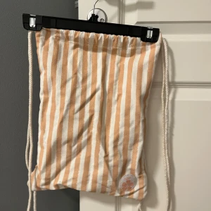 Randig ryggsäck med snören - Säljer en snygg randig ryggsäck i beige/korall rosa och vit med texten 'Aloha' och en blomma på framsidan. 