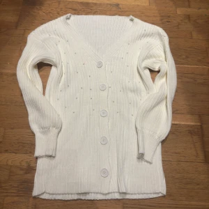 Vit ribbad kofta med knappar - Säljer en stilren vit ribbad kofta med v-ringning och knappar framtill. Perfekt för lager-på-lager och passar till många olika outfits. Koftan har långa ärmar och är både bekväm och mångsidig.