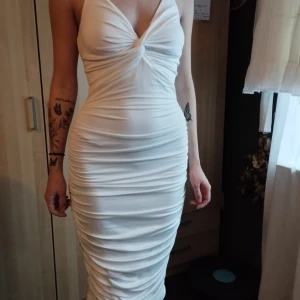 Vit tight klänning - Snygg vit klänning med tight passform och v-ringad design. Klänningen har smala axelband och rynkade detaljer längs sidorna som ger en smickrande silhuett. Perfekt för en elegant kvällslook.