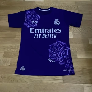 Lila Real Madrid fotbollströja från Adidas - Snygg lila Real Madrid tröja med korta ärmar och coolt ros-mönster. Adidas-logga och 'Emirates Fly Better' tryck på framsidan. Perfekt för fotbollsfans!