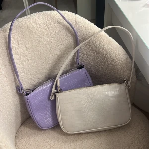 Ljuslila och beige axelväskor  - Två stilrena axelväskor i ljuslila och beige. Båda väskorna har en krokodilpräglad yta och dragkedja upptill. Perfekta för att ge en touch av färg och stil till din outfit. 200 för båda 