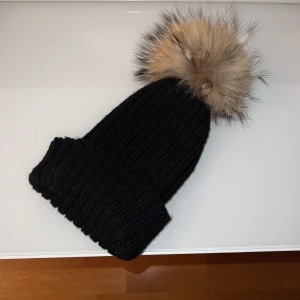Svart stickad mössa med pälsboll - Säljer en superfin svart stickad mössa med en fluffig pälsboll på toppen. Pälsen är äkta. Perfekt för kalla vinterdagar! Mössan är i  bra skick och har en stilren design som passar till allt. Pälsen ger en lyxig touch och gör den extra mysig. Ett måste för lite kyligare väder! 🖤