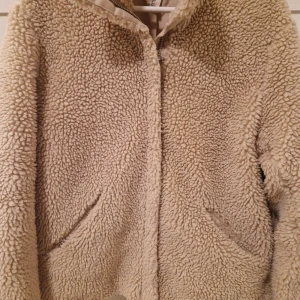 Beige teddyjacka - Mysig beige teddyjacka med dragkedja och långa ärmar. Perfekt för kyliga dagar med sin fluffiga och varma design. Tillverkad av polyester och akryl för extra komfort.