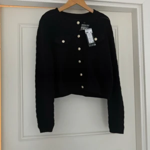 Svart kofta med knappar - Snygg svart kofta med dekorativa knappar och två bröstfickor. Koftan har en ribbad struktur och långa ärmar, perfekt för kyligare dagar. Ett stilrent plagg som passar till många olika outfits.