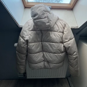 Beige pufferjacka med huva - Säljer en beige pufferjacka med huva och dragkedja framtill. Jackan har långa ärmar och två fickor med dragkedja. Perfekt för kyliga dagar. Sitter som en S