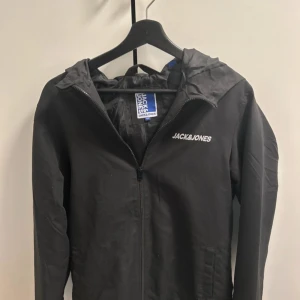 Svart vindjacka från Jack & Jones - Snygg svart vindjacka från Jack & Jones med dragkedja och huva. Perfekt för blåsiga dagar. Jackan har en enkel design med logotyp på bröstet.