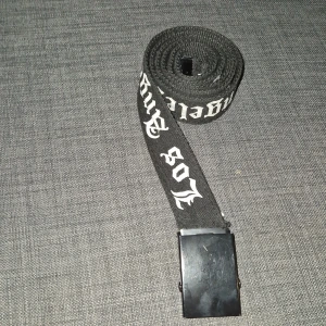 Svart bälte med text -  Svart bälte med vit text 'Los Angeles' i gotisk stil. Bältet har ett stilrent svart spänne och är perfekt för att ge en edgy touch till din outfit.
