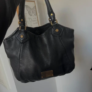 Svart skinnväska från Marc by Marc Jacobs - Snygg svart skinnväska från Marc by Marc Jacobs med gulddetaljer och ett stilrent mönstrat foder. Väskan har två rejäla handtag och en praktisk storlek för vardagsbruk. Perfekt för att bära med sig alla nödvändigheter med stil.