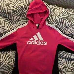 Rosa hoodie från Adidas - Säljer en snygg rosa hoodie från Adidas med vit logga på bröstet. Tröjan har svarta och vita detaljer på ärmarna och är väldigt skön. Helt fläckfri och har använt en gång och säljer pga att den är för kort för mig💗💗💗