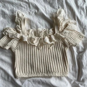 Beige ribbad topp från Zara - Charmig beige ribbad topp från Zara med volangdetaljer och spets vid axlarna. Toppen har en croppad passform och är perfekt för en somrig look. Den är tillverkad i ett mjukt material som ger en romantisk känsla.