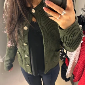 Olivgrön stickad kofta med knappar - Säljer en olivgrön stickad kofta med dekorativa knappar. Koftan har långa ärmar och ribbade detaljer vid ärmslut och nederkant. Perfekt för kyliga dagar! 