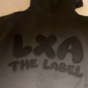 Svart hoodie från LXA The Label - Säljer en svart hoodie från LXA The Label med stor logga på ryggen. Den har en bekväm passform med huva. Perfekt för en avslappnad stil. Mörkgrå/mörkblå