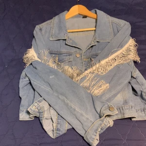 Ljusblå jeansjacka med fransar - Trendig ljusblå jeansjacka med fransdetaljer längs ärmarna och axlarna. Jackan är croppad och har knappar framtill. Perfekt för en avslappnad stil.