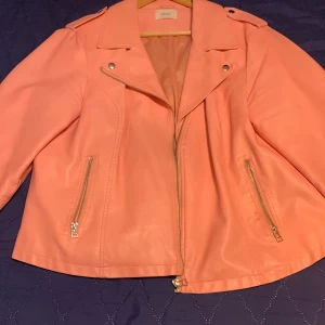 Rosa skinnjacka från Zizzi - Snygg rosa skinnjacka från Zizzi med dragkedja och stiliga detaljer. Jackan har en klassisk bikerstil med långa ärmar och två dragkedjefickor framtill. Perfekt för att ge din outfit en färgklick!