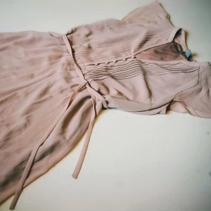 Beige rosa klänning med knappar och knytband - Elegant beige rosa klänning med korta ärmar och knappar framtill. Den har ett knytband i midjan för en smickrande passform. Perfekt för vår och sommar.
