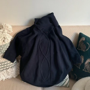 Svart poncho från woolmark - Säljer en svart poncho i ren merinoull från Woolmark. Tröjan har en hög krage och korta ärmar, perfekt för kyligare dagar. Den är mjuk och bekväm, idealisk för lager-på-lager-stil. Passar både till vardags och mer uppklädda tillfällen.