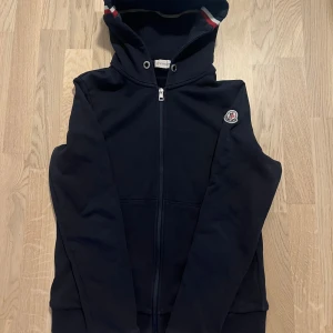 Mörkblå zip hoodie från Moncler - Säljer en mörkblå huvtröja från Moncler med dragkedja och klassisk logga på ärmen. Storlek M men passar mellan S-M. Den är i bra skick MEN har ett lagat hål på ärmen som du ser på sista bilden och därför är priset lite längre.