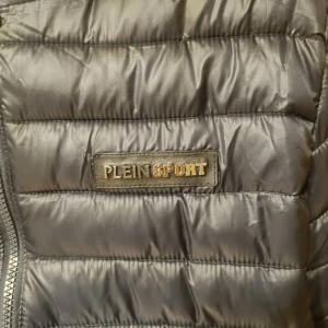 Svart quiltad jacka från Plein Sport - Snygg svart quiltad jacka från Plein Sport med gul logga på ryggen och huvan. Jackan har dragkedja framtill och två sidofickor med dragkedjor. Perfekt för kyligare dagar.